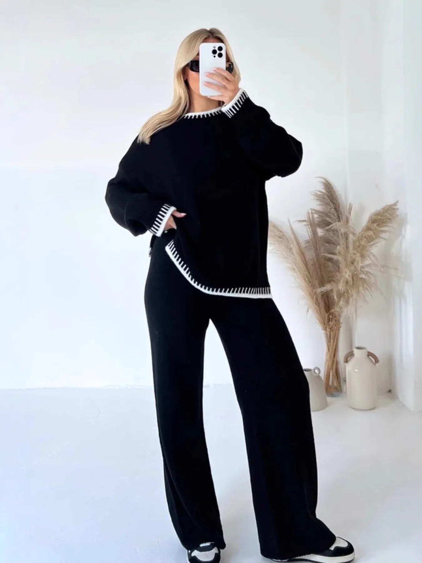 This stylish black sweater set features a round-neck top with contrasting white trim details and matching wide-leg pants. Made from soft knitted material, the set offers ultimate comfort with its relaxed oversized fit. The top showcases decorative white edging on the neckline and hem, while the high-waisted pants provide a flattering silhouette. Perfect for casual outings, lounging, or creating an effortlessly chic look. The coordinated two-piece design ensures a put-together appearance with minimal effort,