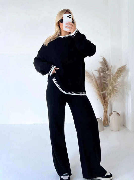 This stylish black sweater set features a round-neck top with contrasting white trim details and matching wide-leg pants. Made from soft knitted material, the set offers ultimate comfort with its relaxed oversized fit. The top showcases decorative white edging on the neckline and hem, while the high-waisted pants provide a flattering silhouette. Perfect for casual outings, lounging, or creating an effortlessly chic look. The coordinated two-piece design ensures a put-together appearance with minimal effort,